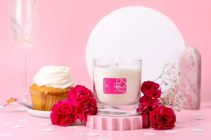 I Choose Love Candle | Aluminate Life in a clear glass holder surrounded by pink carnations, a cupcake with white frosting, and a rose quartz crystal on a soft pink background.