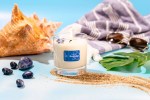 Glass jar candle labeled "I Choose Serenity Candle | Aluminate Life" surrounded by natural elements including seashells, black stones, and green leaves on a woven mat against a soft lavender background, evoking a calm, spa-like wellness atmosphere.