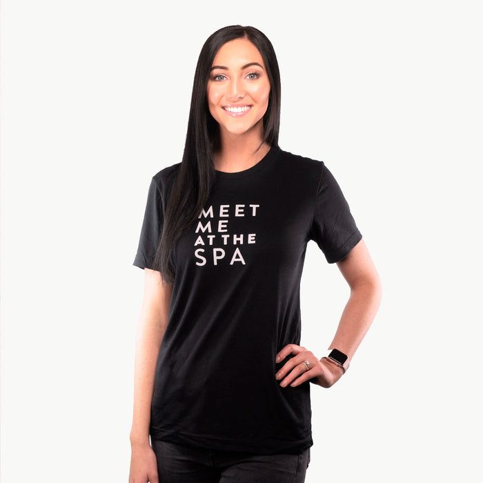 A smiling woman with long dark hair wearing a black Meet Me at the Spa Unisex Crew Neck T-Shirt - Black | Lucky Owl, posing with one hand on her hip against a white background.