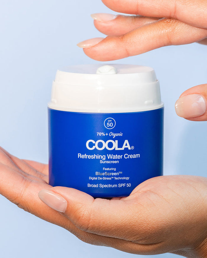 A hand holding a blue jar of Refreshing Water Cream Organic Face Sunscreen SPF 50 | COOLA with a small amount of cream dispensed on top, showcasing skincare and sun protection.