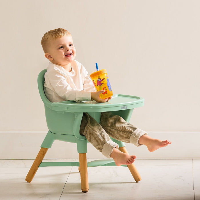 A smiling toddler sits in the Nuby 4-in-1 Multi-Use High Chair | Green Garden, holding a yellow sippy cup with a blue straw. Dressed in a white long-sleeve shirt and beige pants, their bare feet rest on the chair’s tray.