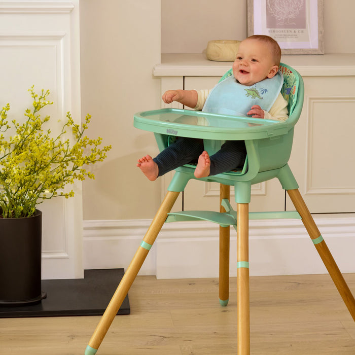A smiling baby sits in the Nuby 4-in-1 Multi-Use High Chair | Green Garden with wooden legs, wearing a blue bib and dark pants, in a cozy room with beige cabinets and a potted plant.