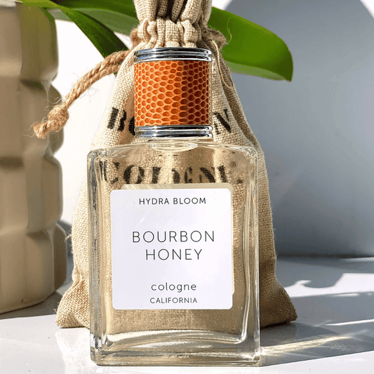Clear glass bottle of Bourbon + Honey Cologne - Unisex Masculine Scent | Hydra Bloom with textured honeycomb-patterned cap, natural light, and burlap pouch in background.