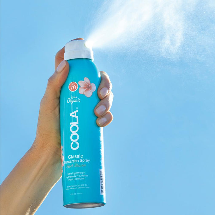 Hand holding and spraying Classic Body Organic Sunscreen Spray SPF 70 | COOLA with a light mist against a clear blue sky background, highlighting skincare and sun protection.