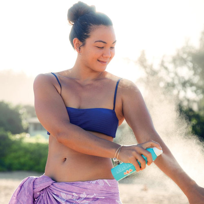 Woman wearing a blue bikini applying Classic Body Organic Sunscreen Spray SPF 70 | COOLA on her arm outdoors, promoting skincare and sun protection.