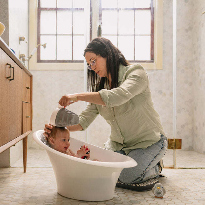 A woman kneels beside a small bathtub, gently pouring water over a baby using the Collapsible Shampoo Rinse Cup. The baby sits in the tub looking up as natural light streams in through the windows.