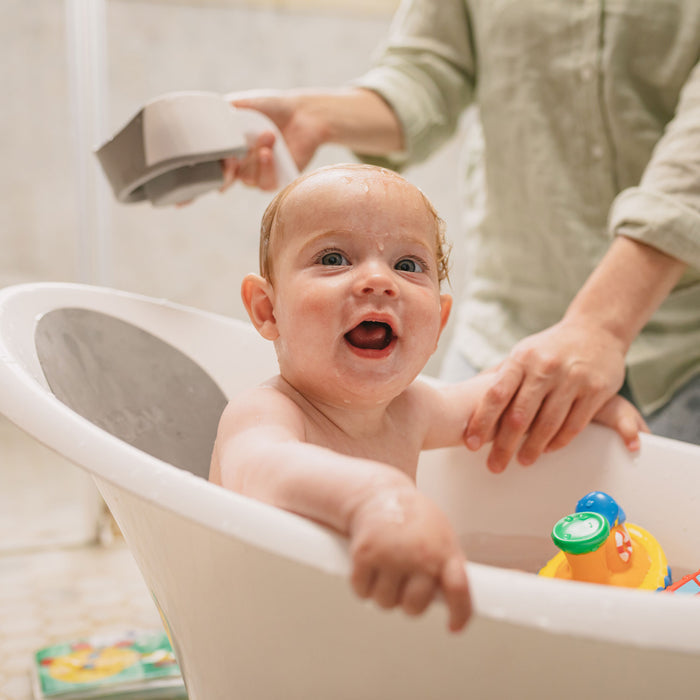 A smiling baby sits in a white tub with colorful toys as an adult uses a handheld shower and the Collapsible Shampoo Rinse Cup for a gentle, tear-free bath. The baby looks up, enjoying the handy rinse cup as a bath accessory.