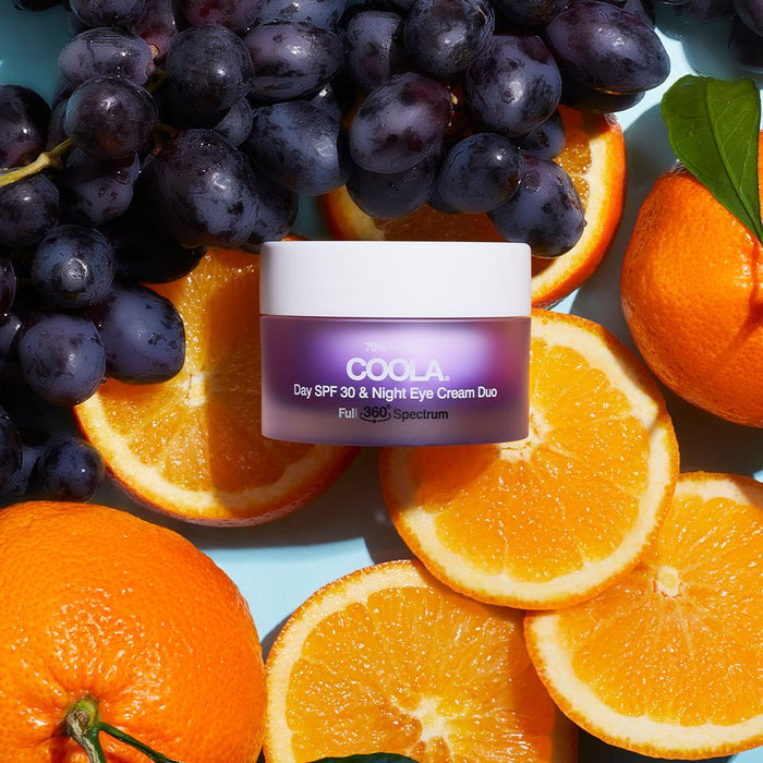 Jar of Full Spectrum 360° Day SPF 30 & Night Organic Eye Cream Duo | COOLA surrounded by fresh oranges and purple grapes, highlighting natural skincare ingredients.