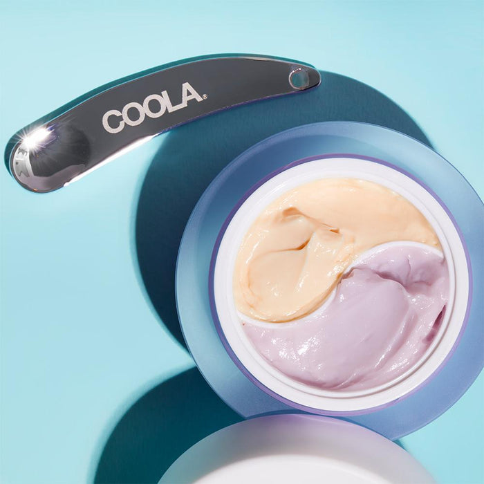 Open jar showing two-tone cream in lavender and beige shades with a metal applicator embossed with COOLA, featuring Full Spectrum 360° Day SPF 30 & Night Organic Eye Cream Duo | COOLA for skincare.