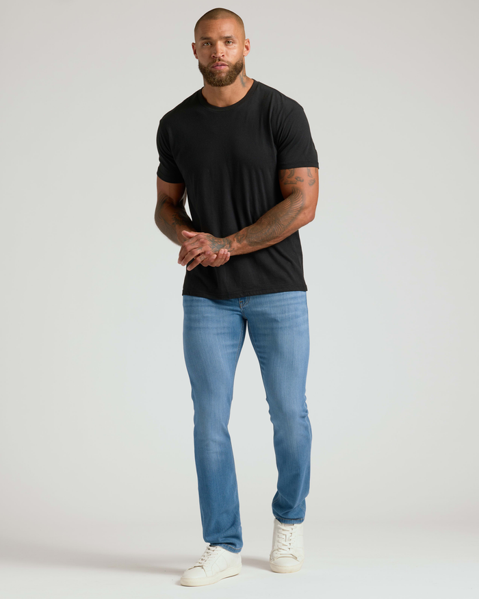 Athletic man wearing light blue Slim Comfort Stretch Jeans 3-Pack paired with a black t-shirt and white sneakers against a plain background.
