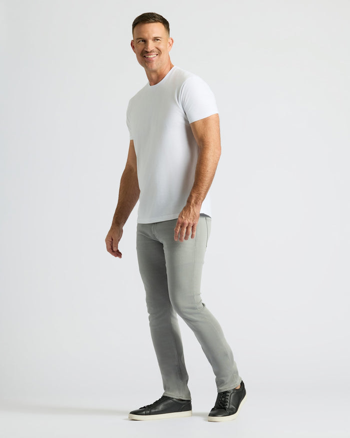 Man wearing Gray Medium Indigo Slim Comfort Stretch Jeans 3-Pack with a white t-shirt and black sneakers, standing against a plain light background in a relaxed pose.