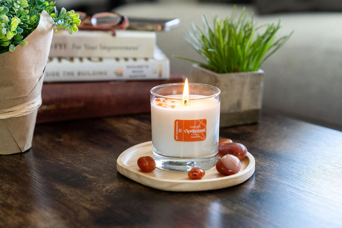 I Choose Optimism Candle | Aluminate Life lit on a wooden tray surrounded by polished stones, with plants and books softly blurred in the background, creating a calm wellness scene.