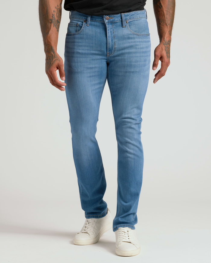 Model wearing light blue Slim Comfort Stretch Jeans 3-Pack in a slim fit with subtle fading, paired with white sneakers against a plain background.