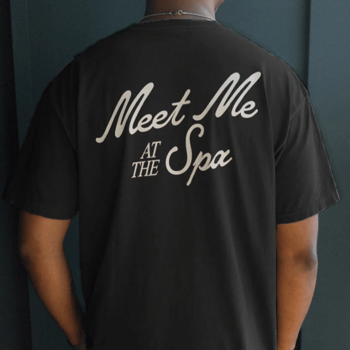 Person wearing a black "Meet Me At The Spa Retro Unisex T-Shirt | Lucky Owl" with white cursive text on the back, promoting a relaxed and wellness-inspired style.