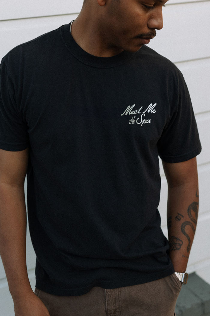 Man wearing a black "Meet Me At The Spa Retro Unisex T-Shirt | Lucky Owl" with white script text on the chest, standing against a white wooden wall.