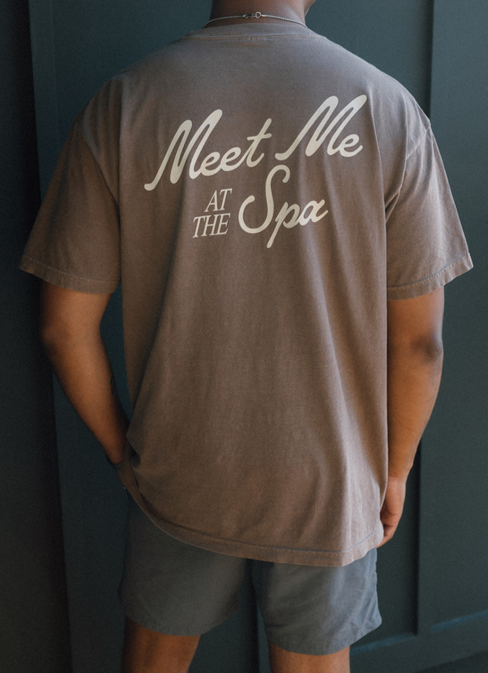 Person wearing a brown Meet Me At The Spa Retro Unisex T-Shirt | Lucky Owl with white text on the back, paired with gray shorts, standing against a dark background.