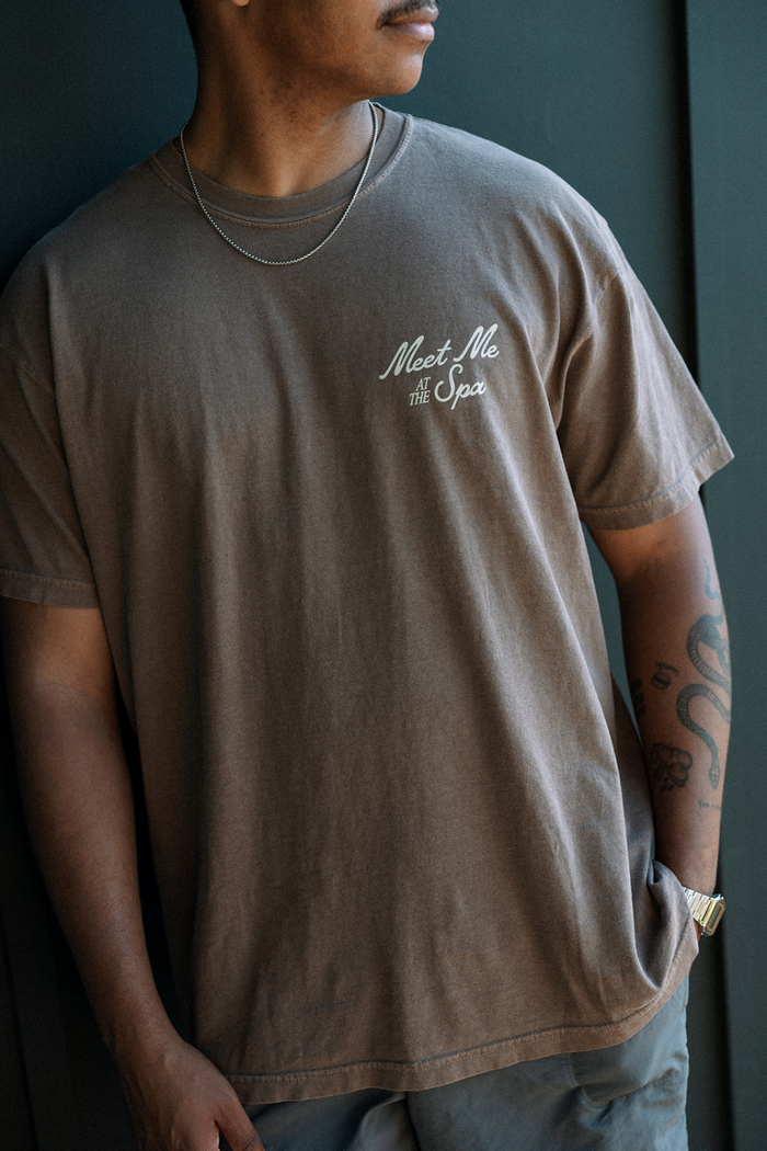 Man wearing a brown Meet Me At The Spa Retro Unisex T-Shirt | Lucky Owl with a small white logo on the chest, paired with a silver chain necklace and casual shorts, standing against a dark background.