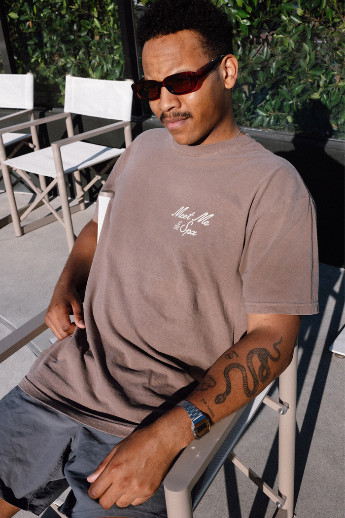 Man wearing sunglasses seated outdoors in a casual setting, dressed in a brown "Meet Me At The Spa Retro Unisex T-Shirt | Lucky Owl" featuring a small white logo on the chest, with visible arm tattoos and a watch.