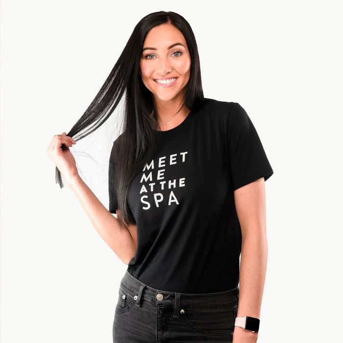 Woman wearing a black Meet Me at the Spa Unisex Crew Neck T-Shirt - Black | Lucky Owl smiling and holding her long dark hair, styled casually with black jeans and a smartwatch.