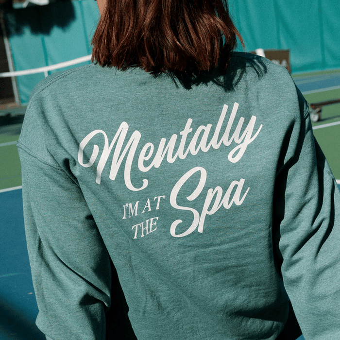 Person wearing a teal sweatshirt with white cursive text reading "Mentally I’m At The Spa" on the back, showcasing the Mentally I’m At The Spa Unisex Crew Neck Sweatshirt | Lucky Owl design.