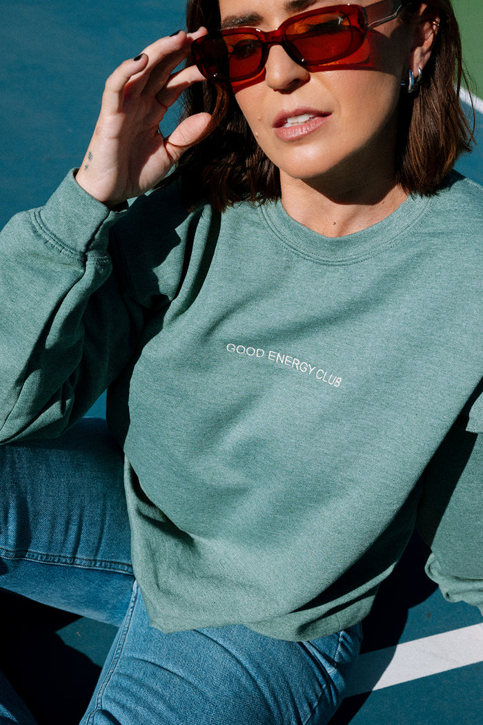 Woman wearing a muted green sweatshirt with "GOOD ENERGY CLUB" printed on the chest, paired with blue jeans and brown sunglasses in natural light, showcasing the Mentally I’m At The Spa Unisex Crew Neck Sweatshirt | Lucky Owl.