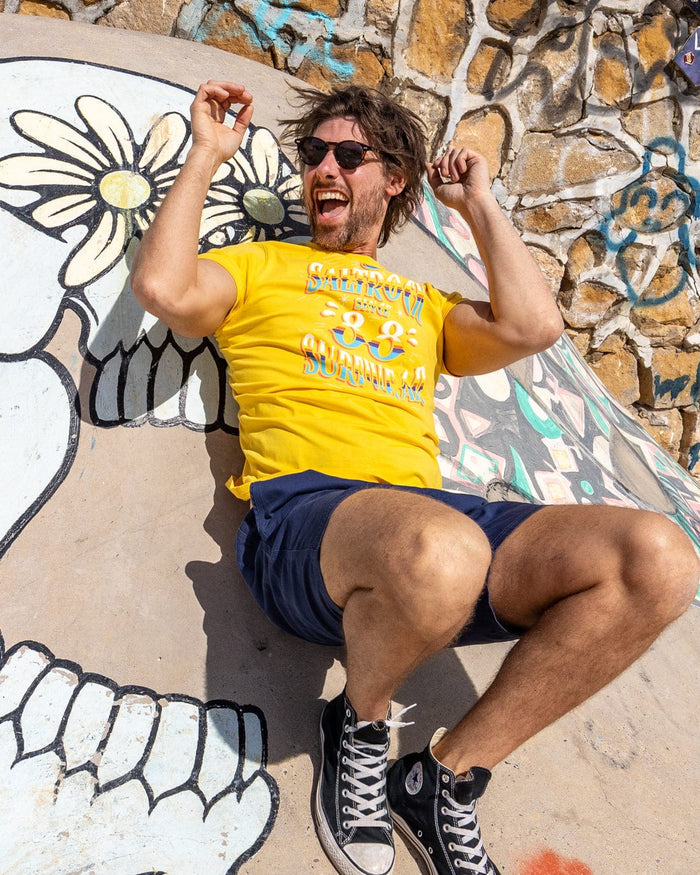 A man in a yellow Mex Logo men's T-shirt by Saltrock and navy shorts sits on a mural-covered concrete surface, smiling with bent legs and raised arms, wearing black Converse sneakers.