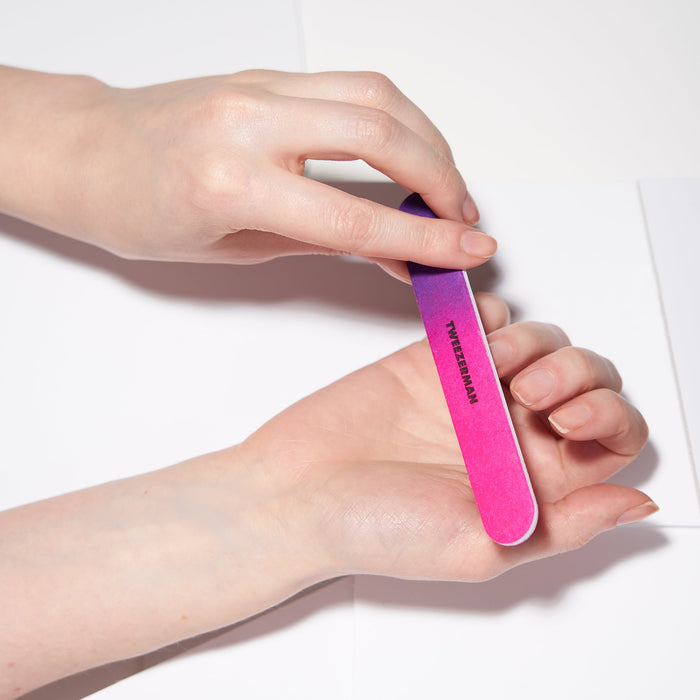 Hands holding and using a brightly colored gradient pink and purple Neon Filemate Set | Tweezerman nail file for gentle nail care and shaping.