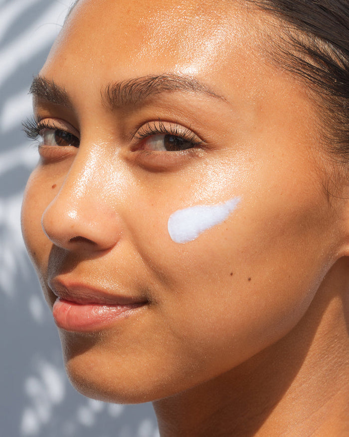 Close-up of a woman with glowing skin and a white cream applied on her cheek, showcasing Refreshing Water Cream Organic Face Sunscreen SPF 50 | COOLA for skincare and sun protection.
