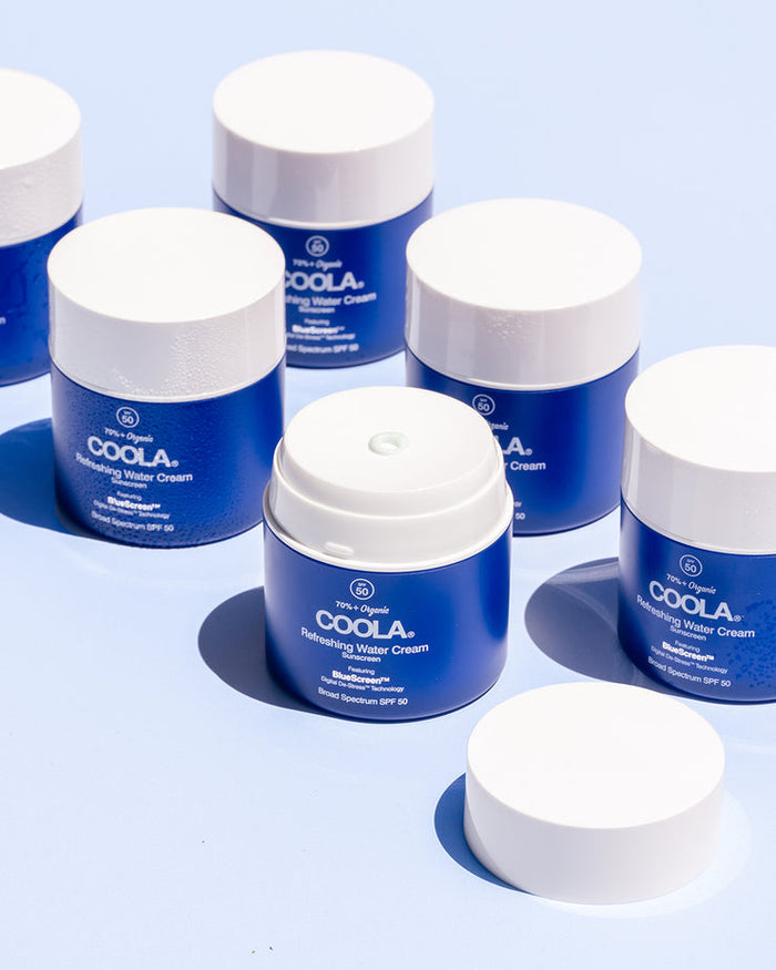 Multiple blue jars with white lids of Refreshing Water Cream Organic Face Sunscreen SPF 50 | COOLA are arranged on a light surface, with one jar open to show the cream dispenser.