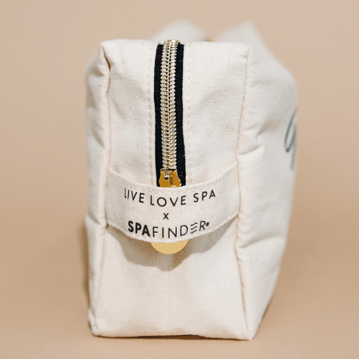 Close-up of a cream-colored canvas travel pouch with a gold zipper, featuring a label that reads "Live Love Spa x Spafinder" for the Spafinder Travel Kit | Live Love Spa.
