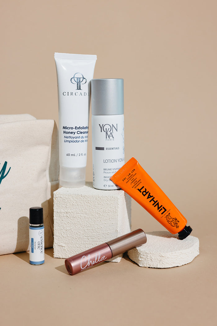 Skincare and body care products including cleanser, lotion, balm, and mascara arranged on textured stone blocks with a fabric bag in the background, showcasing Spafinder Travel Kit | Live Love Spa.