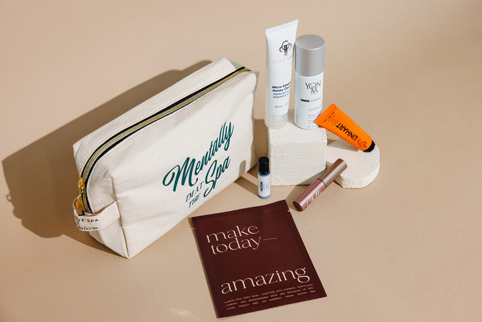 Travel kit with various skincare and wellness products arranged next to a cream-colored canvas pouch labeled "Mentally the Spa," part of the Spafinder Travel Kit | Live Love Spa collection.