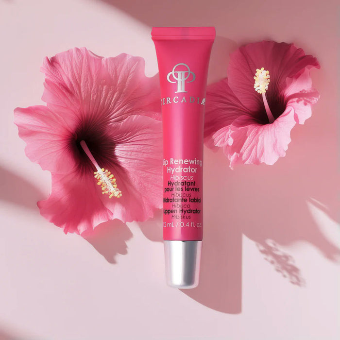 Tube of pink Arcadia Lip Renewing Hydrator placed between two hibiscus flowers on a soft pink background, featured in the Spafinder Travel Kit | Live Love Spa.