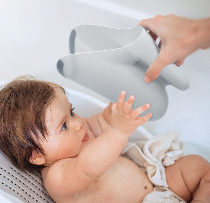 A baby in a bath reaches for a hand holding the Tear-Free Rinse Pail with an easy grip handle, while sitting on a bath support with a towel draped over them.