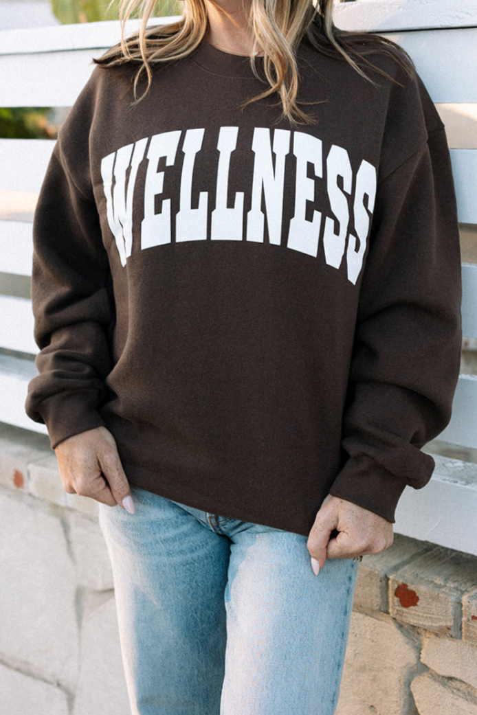 Person wearing a dark brown Wellness Varsity Unisex Crew Neck Sweatshirt | Lucky Owl with large white "WELLNESS" text, paired with light blue jeans, standing outdoors by a white fence.